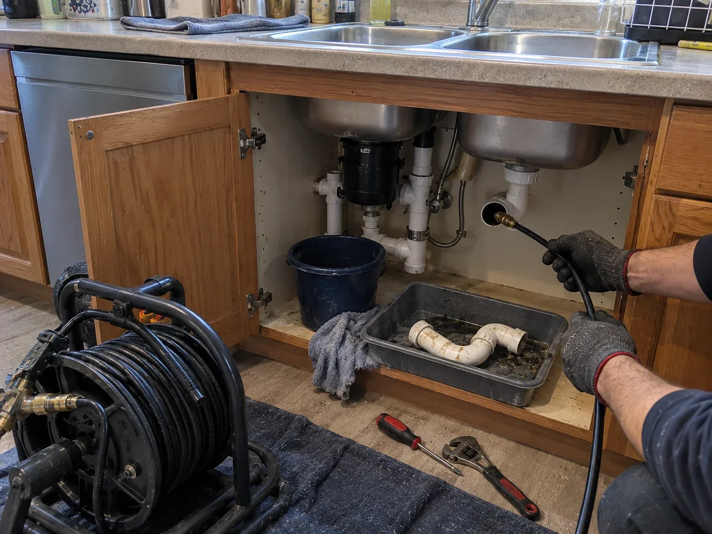 Plumbing technician using a compact jetting hose to clear a kitchen drain beneath a sink in Kitchener